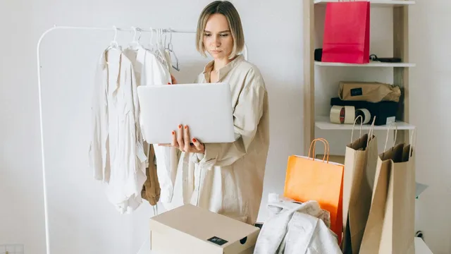 Woman using laptop for online shopping with clothes and bags around her, indoors.